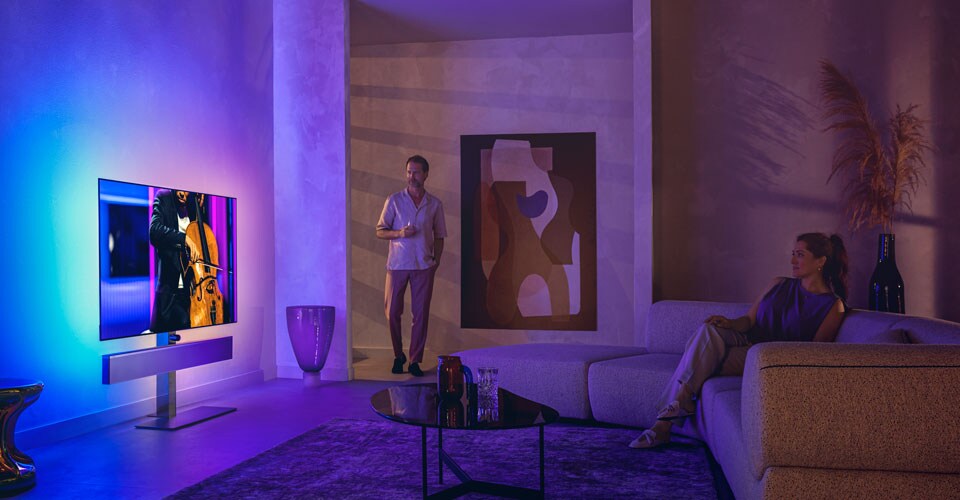 Vyras ir moteris stilingoje svetainėje žiūri „Philips Ambilight“ televizorių, kuriame rodomas violončelės pasirodymas