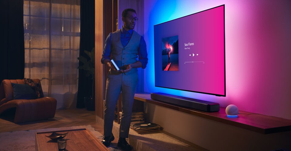 Žmogus modernioje svetainėje šalia „Philips Ambilight“ televizoriaus su muzikos sąsaja ir „Fidelio“ garso juosta