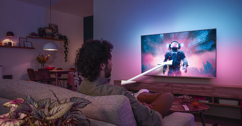 Vyras sėdi ant sofos ir žiūri „Philips Ambilight“ televizorių, kuriame rodomas spalvingas astronauto vaizdas su rodykle, rodančia optimalų žiūrėjimo atstumą