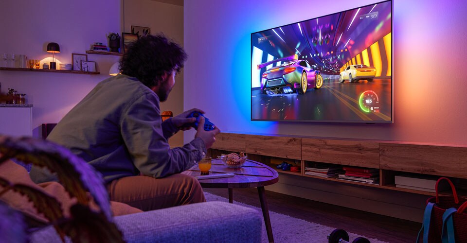 Žmogus sėdi ant sofos ir žaidžia lenktynių vaizdo žaidimą „Philips Ambilight“ televizoriuje