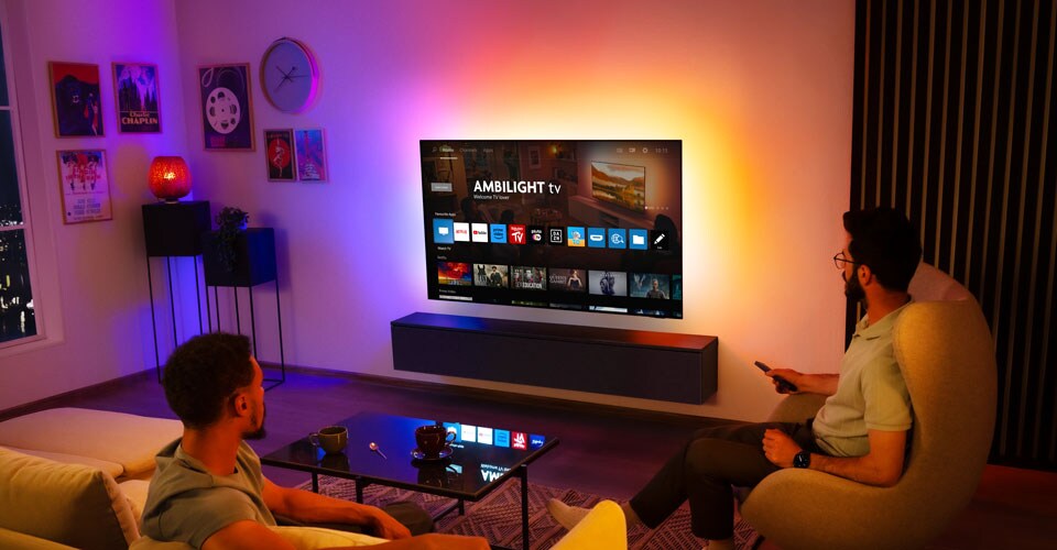 Du vyrai žiūri į prie sienos pritvirtintą „Ambilight“ televizorių svetainėje